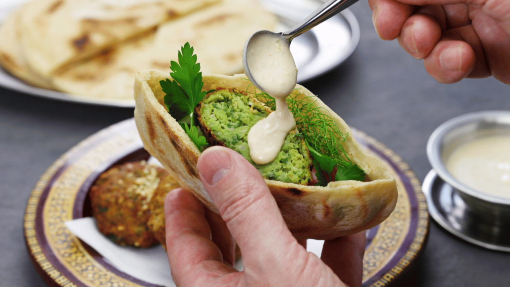 Hand holding open pita bread with Egyptian Falafel inside. Other hand is spooning sauce on top. 