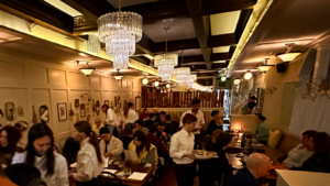 Interior of Perle restaurant, tables full of guests and waitstaff in white shirts walk the dining room under chandeliers.